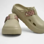 Vista frontal de par de zuecos de plataforma Concept Shoes en color beige arena con correa de patrón monograma rosa sobre fondo blanco.