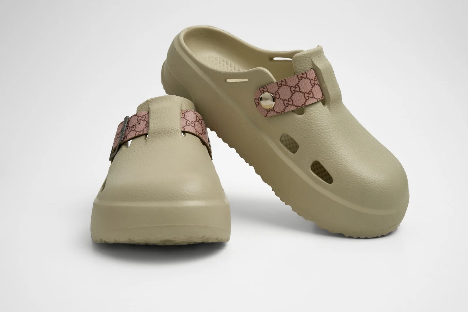 Vista frontal de par de zuecos de plataforma Concept Shoes en color beige arena con correa de patrón monograma rosa sobre fondo blanco.