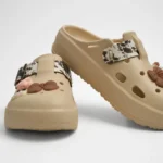 Vista frontal de par de zuecos de plataforma Concept Shoes en color beige topo con correa cowprint y pines de lazo y flor sobre fondo blanco.