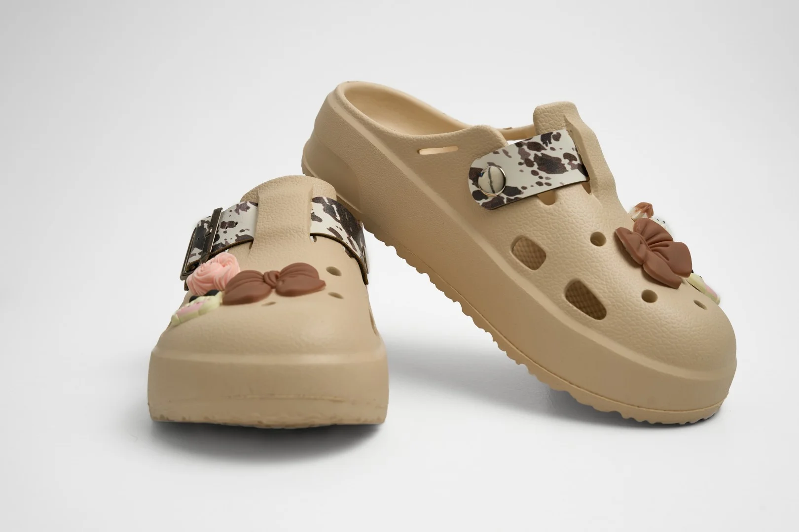 Vista frontal de par de zuecos de plataforma Concept Shoes en color beige topo con correa cowprint y pines de lazo y flor sobre fondo blanco.