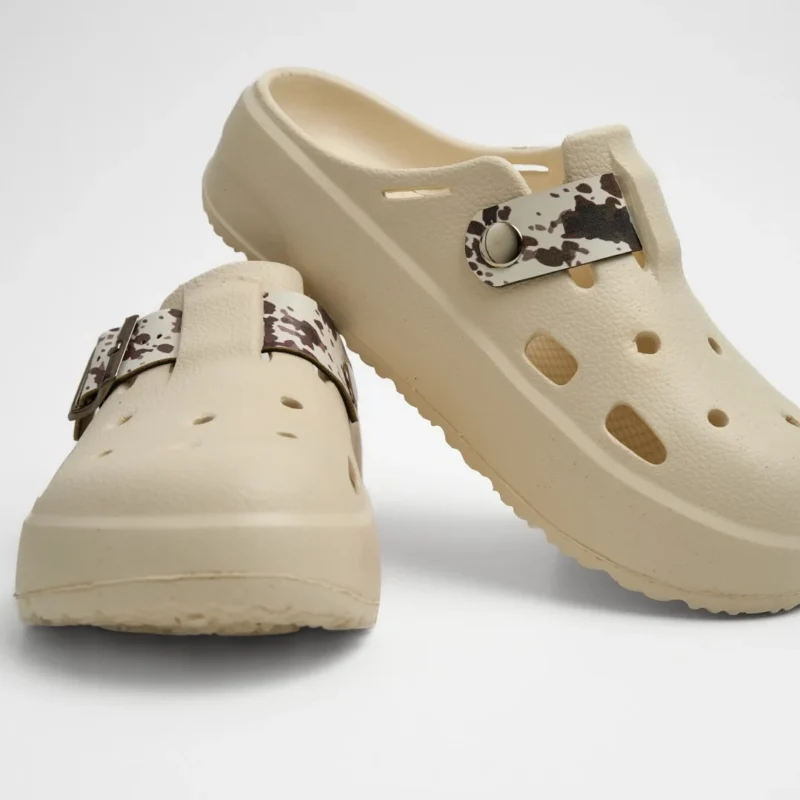 Vista frontal de un par de zuecos de plataforma Concept Shoes en color beige con empeine perforado y correa animal print estilo vaca (cow print) en marrón/blanco sobre fondo blanco.