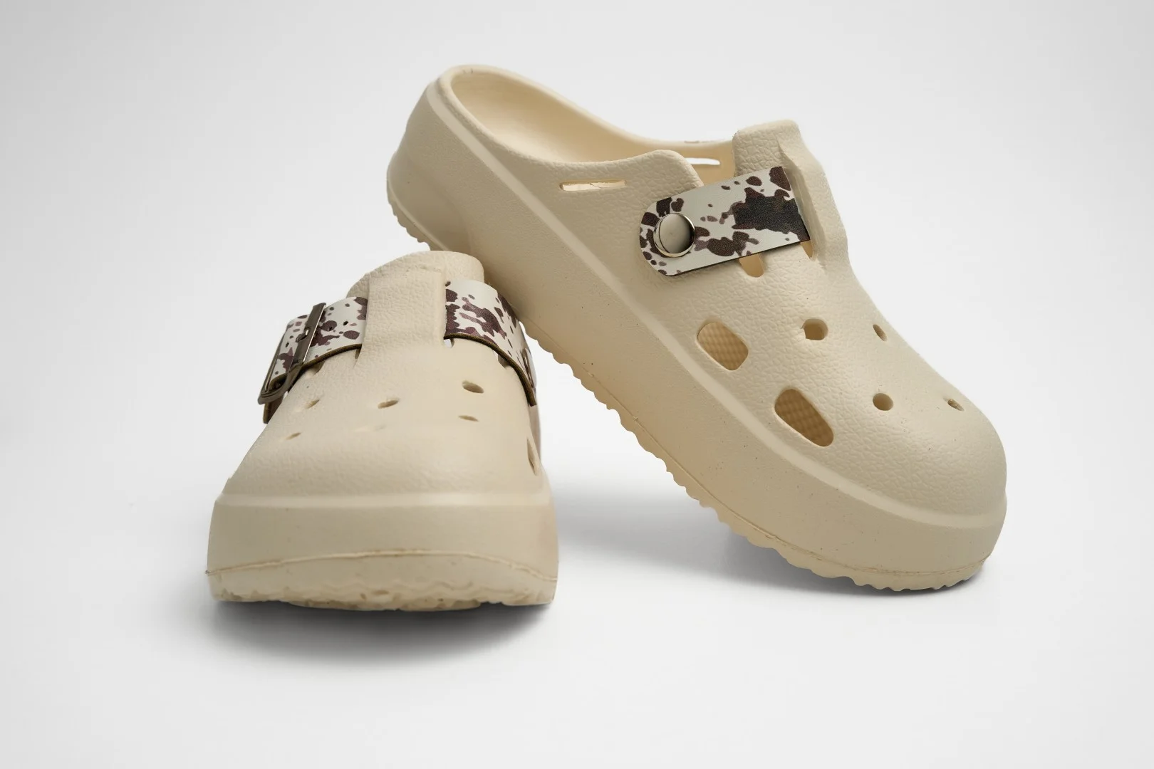 Vista frontal de un par de zuecos de plataforma Concept Shoes en color beige con empeine perforado y correa animal print estilo vaca (cow print) en marrón/blanco sobre fondo blanco.