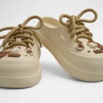 Vista frontal de par de zuecos de plataforma Concept Shoes en color beige topo texturizados con audaces cordones de soga café y dulces charms de postres sobre fondo blanco.