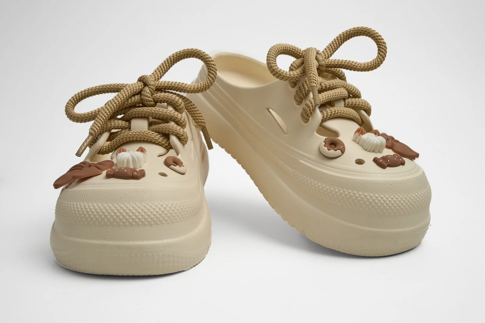 Vista frontal de par de zuecos de plataforma Concept Shoes en color beige topo texturizados con audaces cordones de soga café y dulces charms de postres sobre fondo blanco.