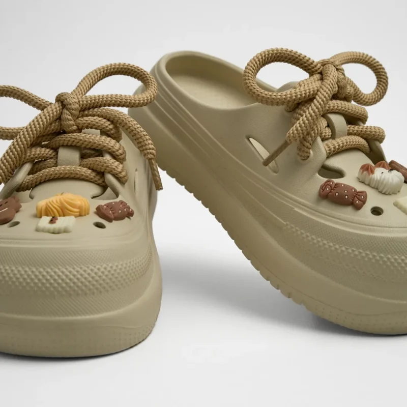 Vista frontal de un par de zuecos de plataforma Concept Shoes en color caqui texturizados con audaces cordones de soga café y charms de postres de chocolate sobre fondo blanco.