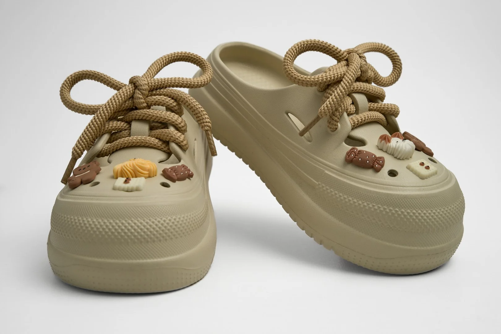 Vista frontal de un par de zuecos de plataforma Concept Shoes en color caqui texturizados con audaces cordones de soga café y charms de postres de chocolate sobre fondo blanco.